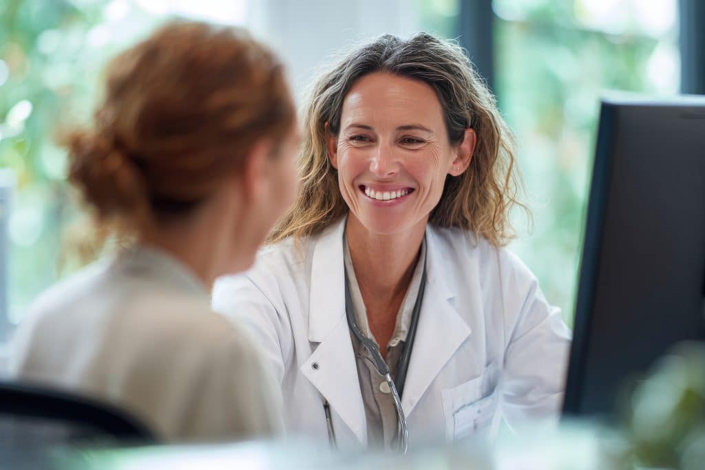 Smiling female doctor wearing a white coat talks to a patient in a bright clinic. The scene suggests a friendly patient consultation.