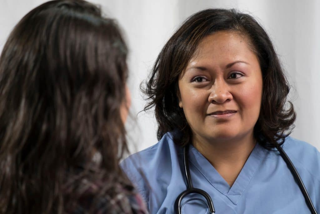 Healthcare worker in light blue scrubs with a stethoscope, smiling at a patient off-camera in a clinical setting