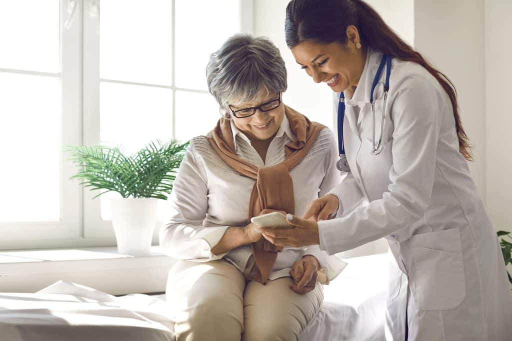 Female doctor in white coat with stethoscope assists an older woman seated by a window, reviewing something on a smartphone.
