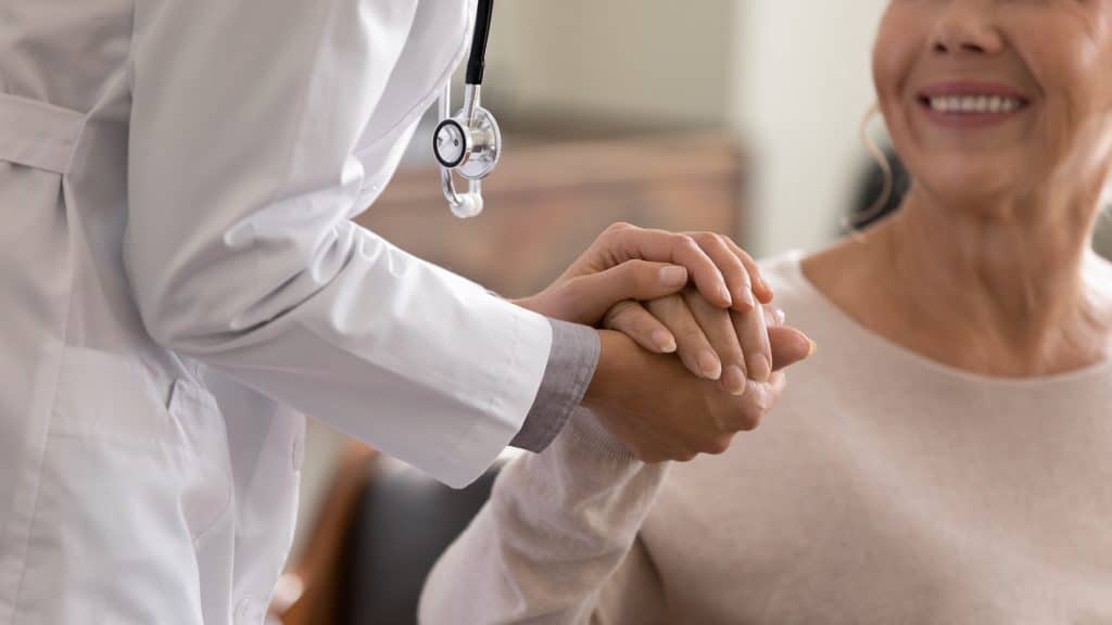 Doctor in a white coat gently holding an elderly patient's hands in a reassuring gesture.