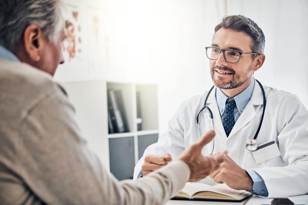 Doctor in a white coat and glasses smiles while shaking hands with a patient across a desk in a clinic, a stethoscope around his neck.
