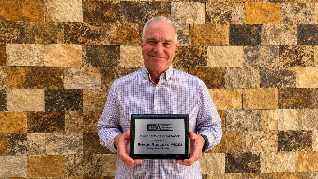Man in a checkered shirt smiling while holding a metal plaque in front of a stone wall; the plaque reads '2025 Chairman's Circle Award'.