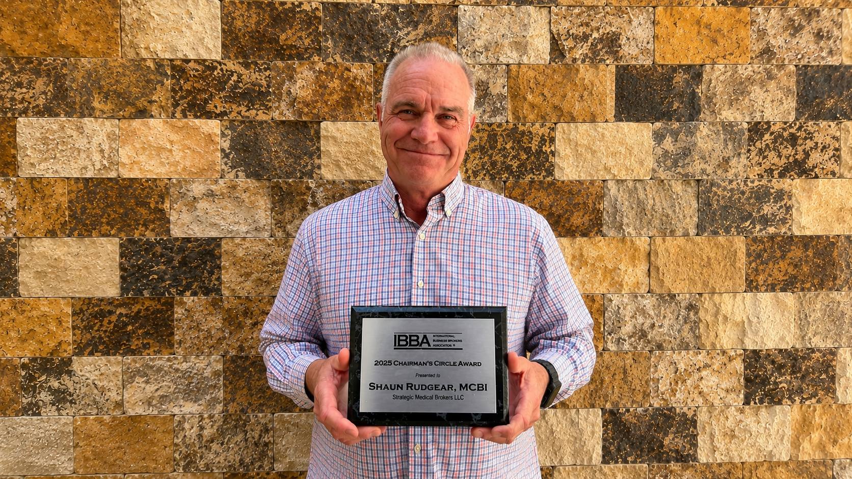 Man in a checkered shirt smiling while holding a metal plaque in front of a stone wall; the plaque reads '2025 Chairman's Circle Award'.