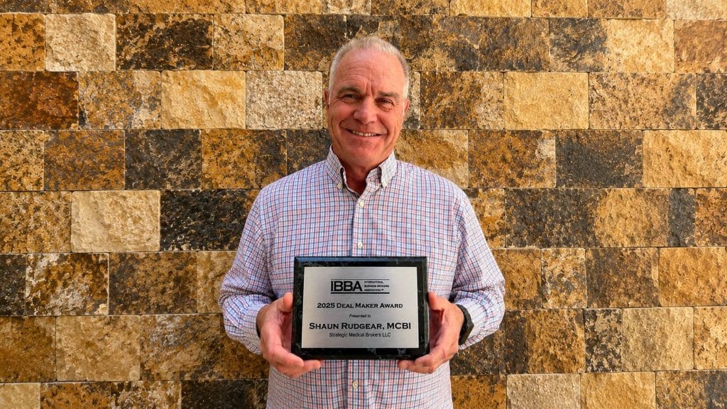 Smiling man in a checked shirt holding a framed award plaque in front of a beige stone wall