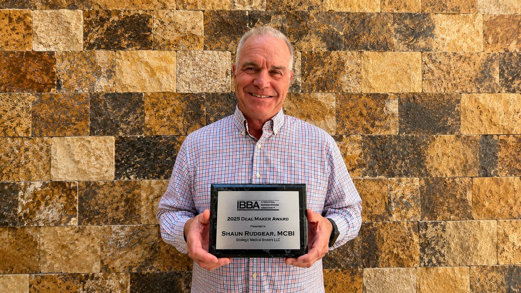 Smiling man in a checked shirt holding a framed award plaque in front of a beige stone wall