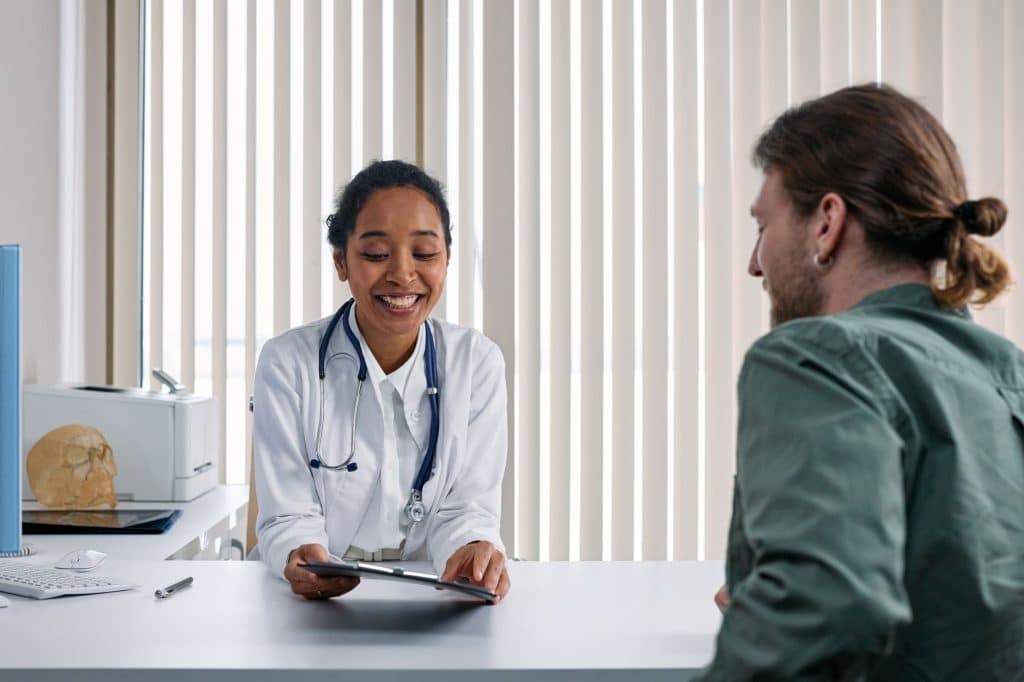 Smiling doctor in a white coat sits at a desk showing a tablet to a patient in a green shirt, during a medical consultation.