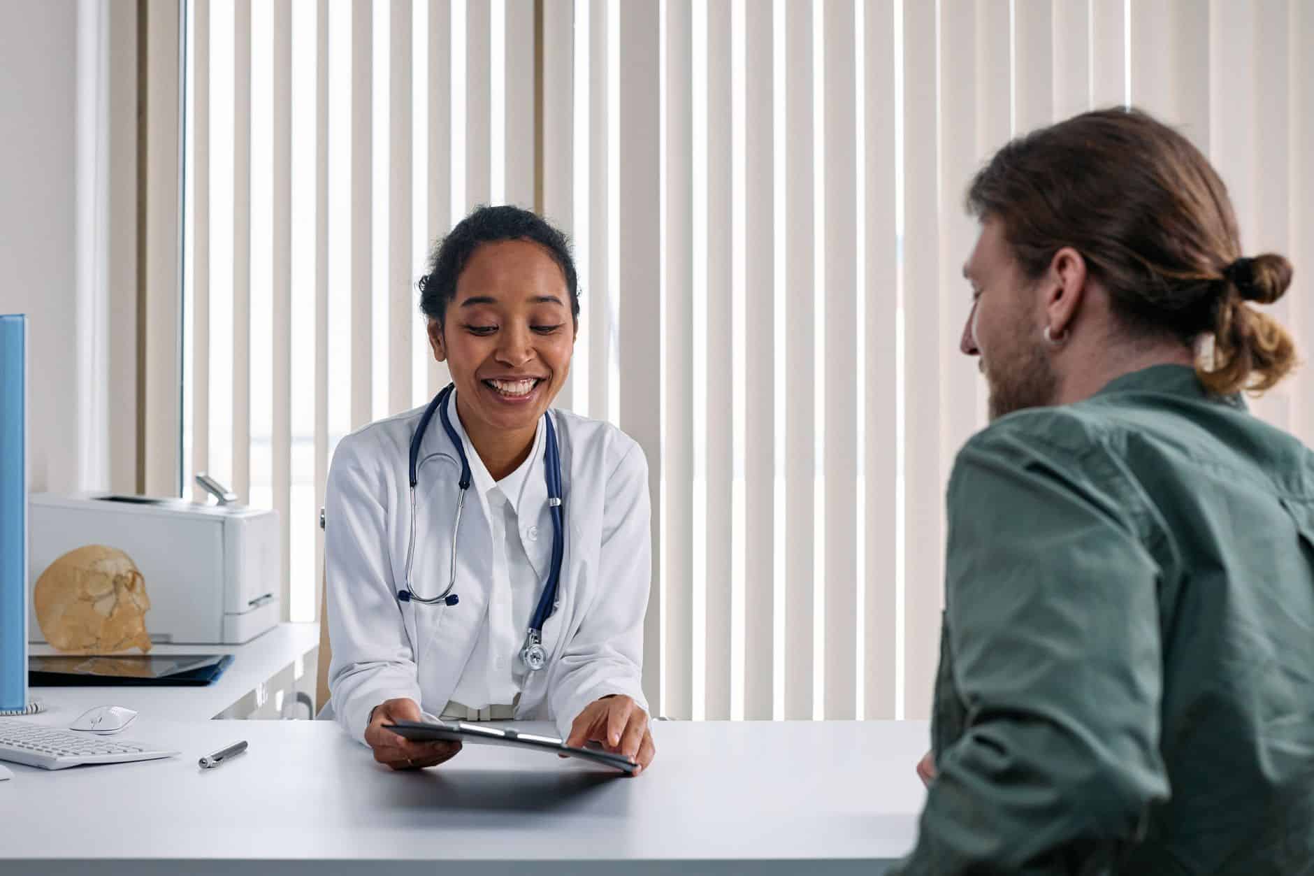 Smiling doctor in a white coat sits at a desk showing a tablet to a patient in a green shirt, during a medical consultation.