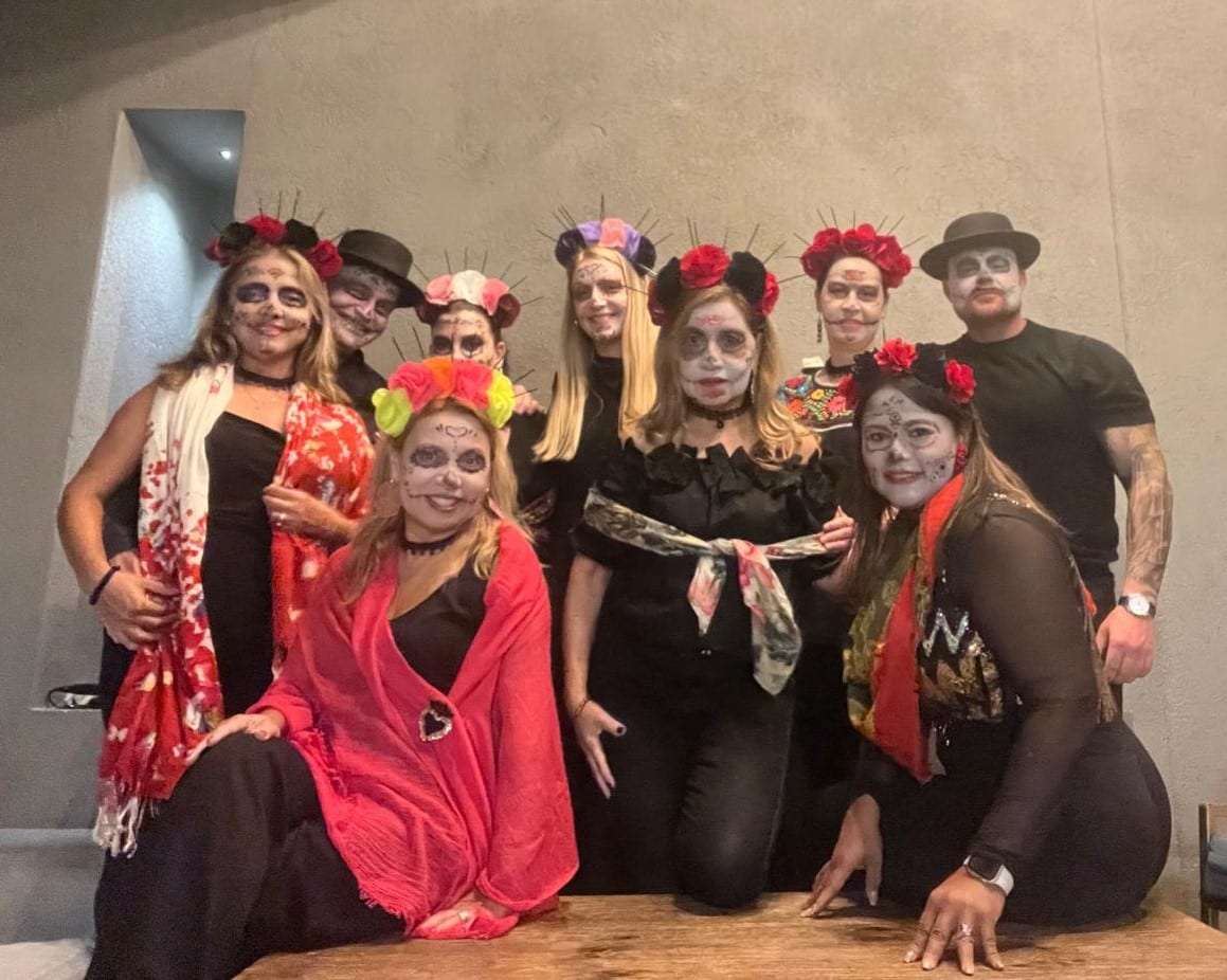 Nine people in Day of the Dead makeup and flower crowns posing together at a party, colorful headpieces and black outfits.
