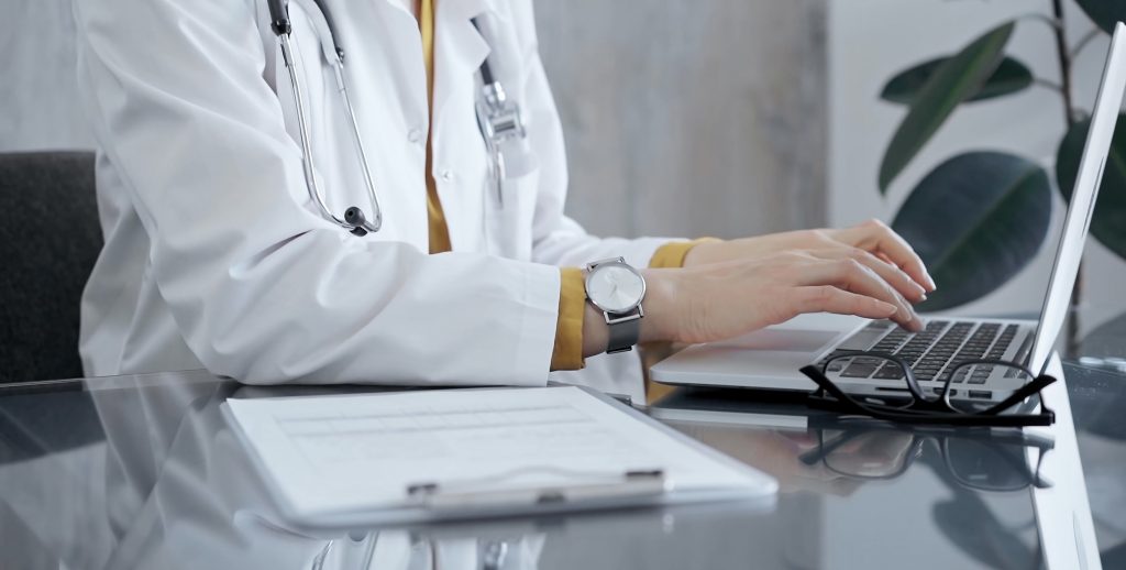 Doctor woman wearing white medical coat over a yellow blouse is working on laptop with stethoscope on the glass desk. Medicine and Health care.