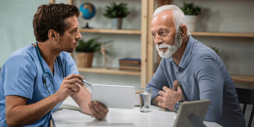 Doctor in scrubs shows a tablet to an elderly man during a medical consultation at a table.