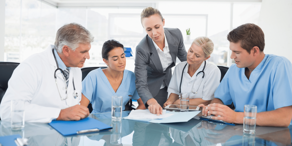 A group of healthcare professionals and a businesswoman discuss documents at a conference table in a bright office.